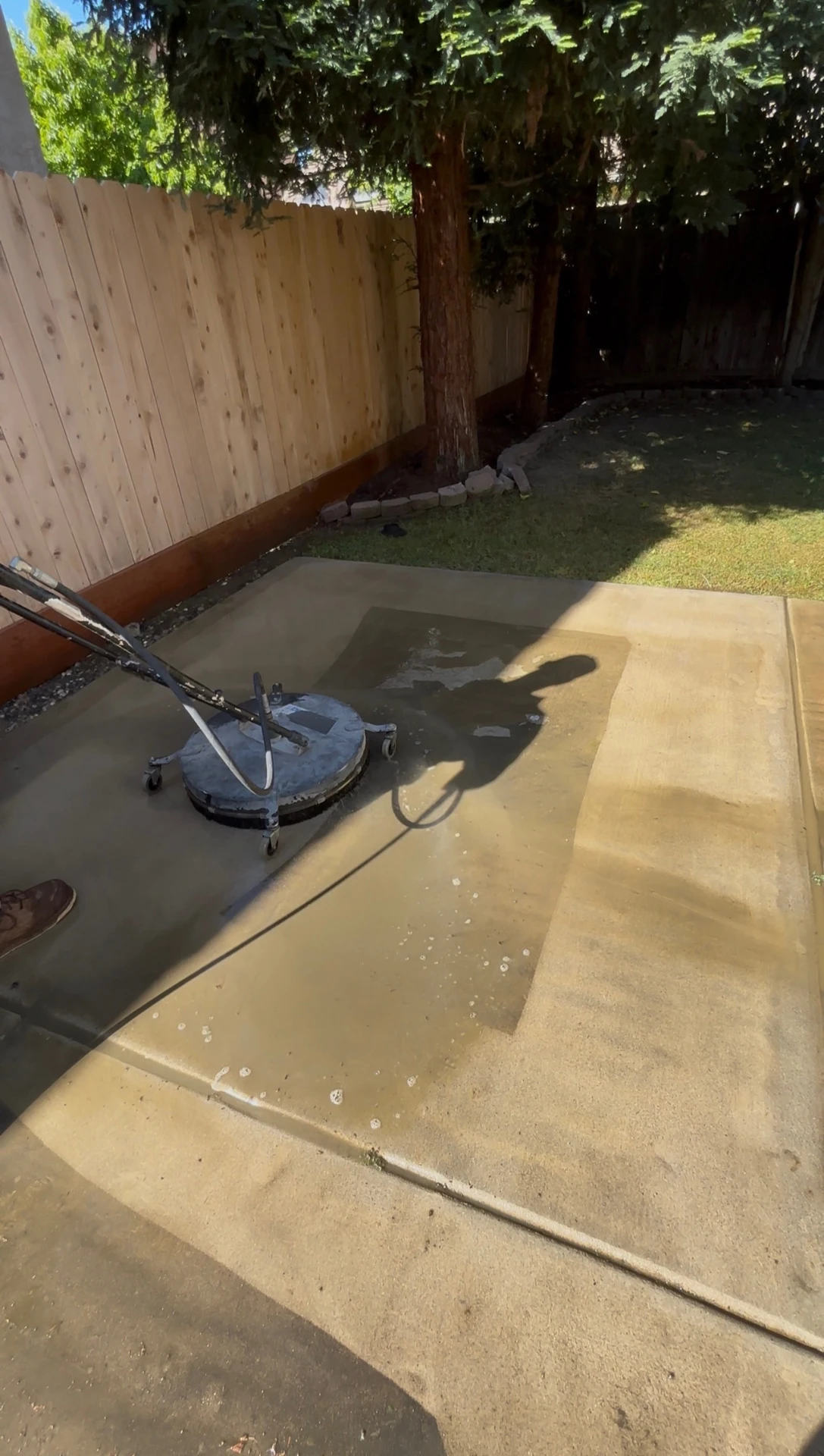 A surface cleaner mid-pass on a backyard concrete patio, with a sharp line between the cleaned and dirty halves of the slab